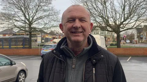 A man with a shaven head is wearing a black coat and a grey fleece while standing in a car park.