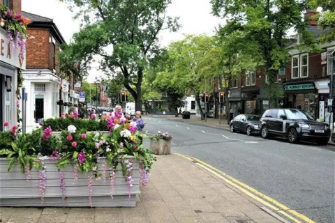 Geograph/G Laird Boutique shops, cafes and bars line a road through the village with floral displays in the foreground. There are a couple of cars parked on the street.