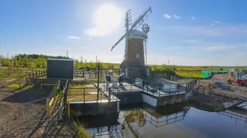 Water Management Alliance The sun can be seen behind a 20th century windpump in the Norfolk Broads, with a new electric pumping station visible in the foreground