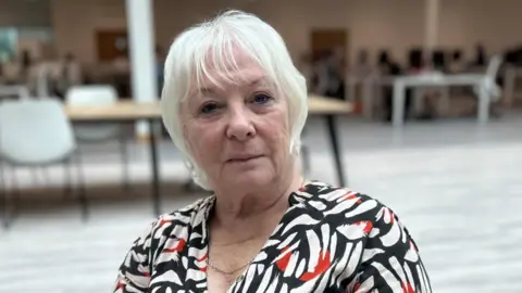 Danielle Stone with medium-length white hair, wearing a top with a white, black and red pattern. She is sitting in a large room with tables and plastic chairs behind her.