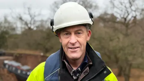 A man is wearing a white hard hat and wearing a yellow fluorescent jacket. a canal boat in the bottom of a large hole is blurred in the background