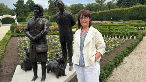 Brenda in front of statue wearing white trousers, a navy and white shirt and a cream jacket.   She has brown hair and black glasses and a pearl drop down earring.   She is smiling with her hands in her pockets.