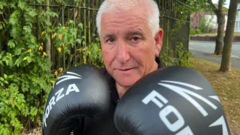 A man in a black T shirt is stood in front of a tree lined fence and looking directly into the camera. He is wearing boxing gloves which are raised to his chin in a fighting pose.