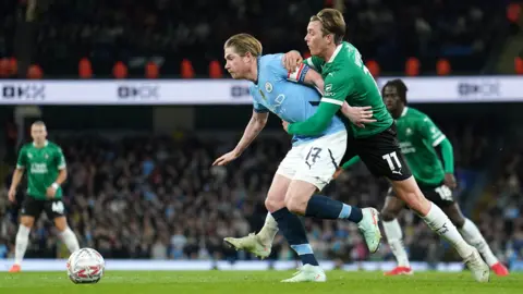 PA Media Manchester City captain Kevin de Bruyne and Plymouth Argyle midfielder Callum Wright tussle while fighting to get the ball in the sides' FA Cup fifth round clash at the Etihad Stadium. De Bruyne has a white and red captain's armband on his left arm. Wright is standing behind De Bruyne and is holding onto the City player.