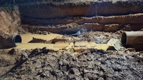 Southern Water Two pipes in a muddy trench. There is cloudy, brown water around and inside one end of pipe. Lots of footprints are visible in the mud.
