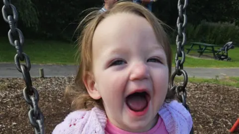 Story family A head and shoulders picture of Lyla - a two-year-old girl. She is on a swing in a children's park. She has short brown hair and is smiling broadly at the camera. She is wearing a pink cardigan and pink top.