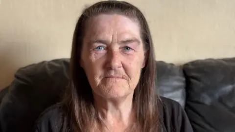 A woman with brown hair in a black top sat on a sofa poses for a photo.
