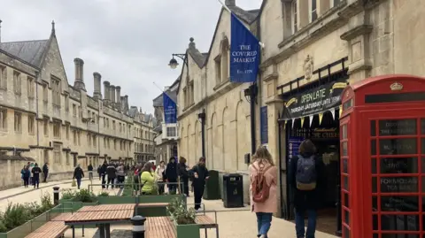 Oxford City Council A street with old buildings on either side stretched into the distance and curves to the right. There are various people dotted around, mostly wearing wintry clothing. Two blue flags jutting out of the wall of a building on the right side of the road read 'The Covered Market est. 1974'. The entrance to the market sits under a banner with 'Open Late' written on it. Two people with their backs turned, both wearing backpacks, appear to be making their way inside. 