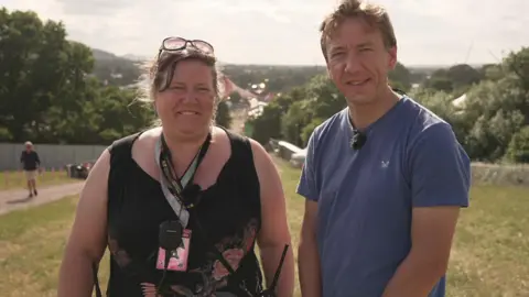 Gareth Churcher and Jade Dunbar standing outdoors with a scenic background of trees and a distant view of a town or festival. Jade Dunbar wears sunglasses on their head, a black sleeveless top, and a lanyard. Gareth Churcher wears a blue t-shirt.