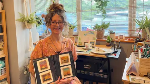 PA Media A woman wearing a pink patterned dress and glasses, sitting in an artist's studio and holding up a book with four tiny paintings in it, painted onto teabags. There's a desk and a window behind her, with plants on the window sill.