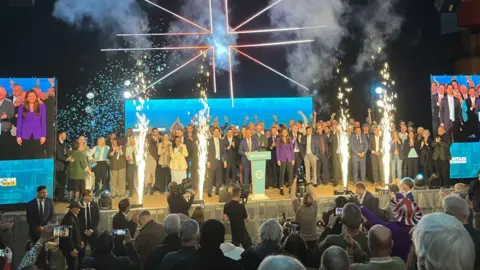A big crowd of people on a stage, with Nigel Farage in the middle, all wearing suits and formal wear. There are four big sparklers at the front of the stage and an audience.