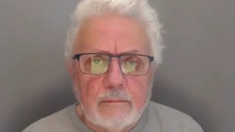 Mugshot of Colin Finch. He has a neat trimmed white beard and short thick white hair. He is wearing dark-framed glasses.