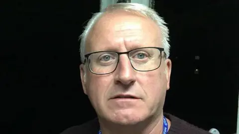 Close up of Glenn, who has white hair and black-rimmed glasses. He is standing or sitting inside with a black background behind him.