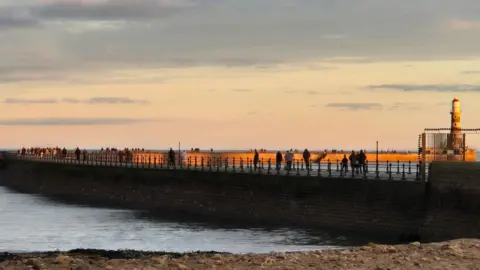 Groups of people walking along Roker Pier on Saturday around sunset, photographed from the beach.