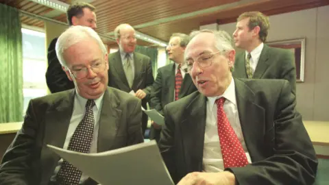 PA Media Jim Wallace and Donald Dewar, both in suits with shirt and tie, sitting side by side looking at a document. They appear to be chatting, and amused.