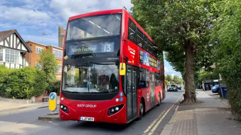 A red London bus is driving along a tree-lined suburban street. There are mid 20th Century houses on both sides of the road. 
