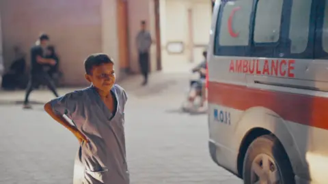 Zakaria wore her blue scrub, as she is standing next to a parked ambulance outside the Al-Aksa Hospital. He has his hands on his hips and looking at the front of the vehicle, he is smiling.