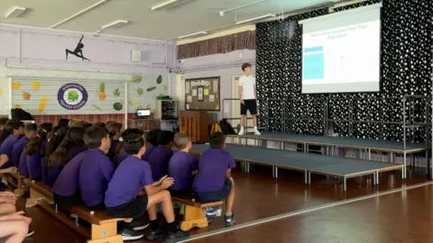 Mathieu is standing on a stage in a school hall in front of a projector screen. He is wearing a white top and black shorts. Pupils sit watching him on wooden benched in purple polo shirts.