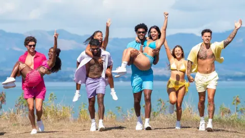 L-R Love Island finalists Nicolas "Nic" Vansteenberghe, Olandria Carthen, Bryan Arenales, Amaya Espinal, Chris Seeley, Huda Mustafa, Iris Kendall, Jose "Pepe" Garcia-Gonzalez. The couples are pictured wearing matching coloured outfits while walking along the coast on a sunny day, the sea and mountains behind them. Nic, Bryan and Chris all carry their partners while Iris and Jose walk hand in hand.