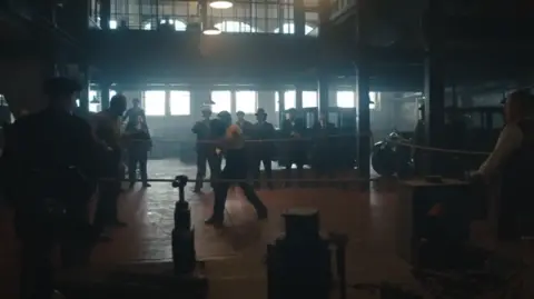 A group of men in suits stand around a makeshift boxing ring in the middle of an old industrial building.