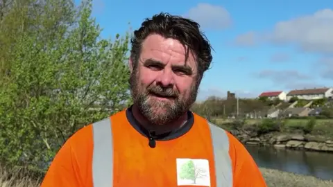 Pat Carr. He has short black hair and a beard. He is wearing a orange high-viz jumper and is speaking to an interviewer out of view. He is stood by the banks of the River Derwent in Workington.