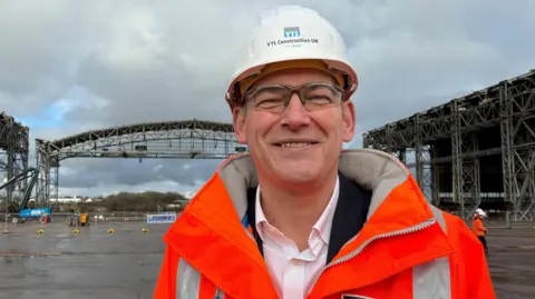 John is wearing fluorescent orange PPE with a white hardhat and safety goggles. He is smiling at the camera and standing in front of the Brabazon hangars.