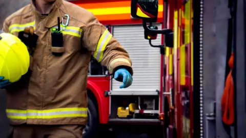 Stock image of firefighter in brown jacket with bright yellow visibility strips and a bright yellow helmet walking alongside two fire engines. 