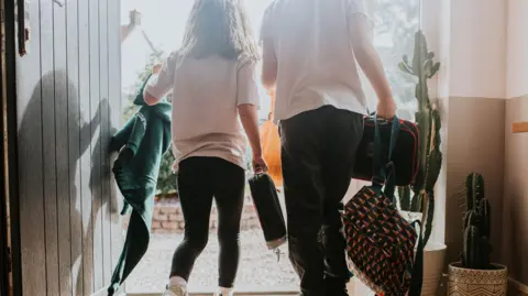 Getty Images Stock picture of the back of two children leaving a house. They are wearing school uniform and carrying bags.