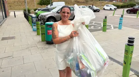 Martin Heath/BBC Sandra McCluskey standing on a pavement outside a supermarket holds a large transparent sack filled with recyclable items, including plastic bottles, cartons and packaging. She is wearing a light-coloured sleeveless outfit and has visible tattoos on both arms and one leg. Behind her are recycling bins, a row of green and blue bollards, parked cars and a supermarket car park. The photo was taken outdoors in daylight, with dry grass visible on a bank in the background.