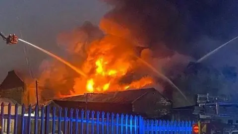 West Yorkshire Fire & Rescue Service A large fire at a warehouse building, with several aerial platforms being used by the fire service. Jets of water are being sprayed on the flames from three directions, with the fire burning brightly. 