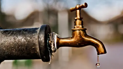 An old looking water pipe with a copper tap on the end. There is a single drip hanging out of the tap.