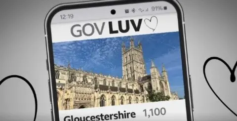 A screenshot of the video used in the article, showing a graphic of a mobile phone with an app that is supposed to look like Tinder. It has the title 'Gov Luv' across the top and a picture of Gloucester Cathedral. It is set against a grey background with black hearts on it. 