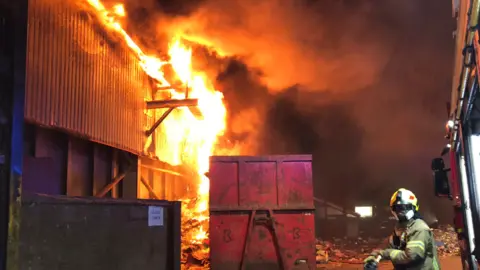 Nottinghamshire Fire and Rescue Service A fire in consuming an industiral building with a firefighter in the foreground