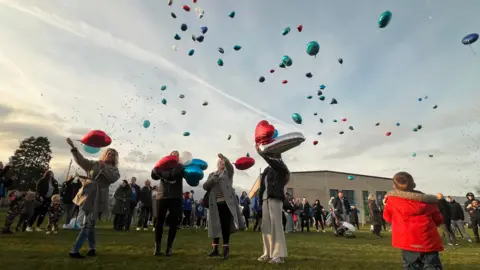 A large crowd gathers in the field of a community centre as they let balloons - mostly red, silver, and blue - into the sky at dusk on a February evening