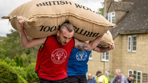 A young man with a red T-shirt and 'Tetbury Woolsack Races' written across it carries a large sack up a hill as part of the Tetbury woolsack races. Behind him is another man with a blue T-shirt with the same logo.
