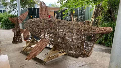Falmouth University The Basket Shark in an outdoors area next to shipping containers during its build phase. The sculpture is mid-build with its tail placed on the floor.