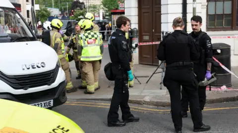 Getty Images Police officers stood outside the corner of a bank which has been closed and there are also fire crews and a large white van parked close by, along with red and white tape blocking off the entrance to the bank, which is a light grey stone building with dark oak doors.