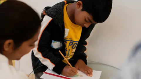 A child drawing a picture on a piece of paper with a pencil, watched by an adult woman