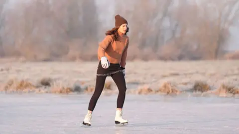 PA Media A woman skating at speed on a frozen field at Upware, Cambridgeshire. She is wearing a brown bobble hat and leggings, an orange jumper and white padded gloves and ice skating boots. Behind her is the field edge and eyond it wintery trees