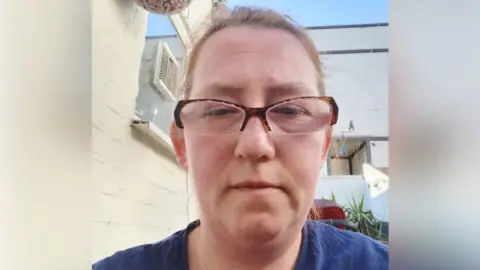 Northamptonshire Police Marta Bednarczky with short brown hair and brown glasses, looking at the camera and wearing a blue top. She is in front of a white building.