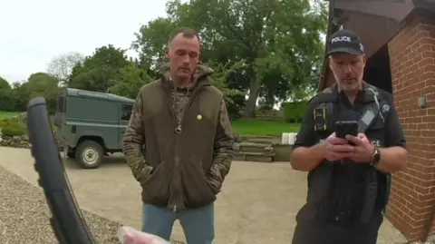 Lincolnshire Police A still from body-worn police footage. A man in a green shirt and jacket with his hand in his pockets has his eyes closed. A police officer is standing next to him typing into his phone. A small green truck and trees can be seen in the background.