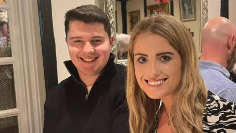 Zak White A young man and woman sit in a pub or restaurant, smiling at the camera. He wears a high-necked zip-up fleece, while the woman wears a top with a black and white pattern reminiscent of an animal print.