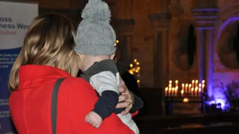 A woman in a red coat stands looking at a display of candles in Lincoln Cathedral.  She is holding a baby in a bobble hat close to her.  Both have their backs to the camera.