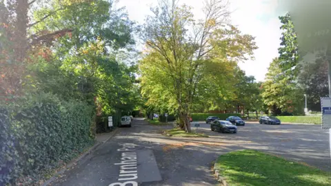Google A screenshot from Google street view showing a car park and a road lined with trees on a sunny day.