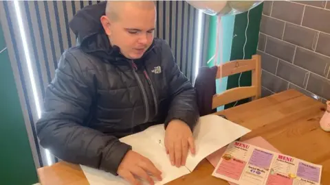The image shows a bald man in a black coat using his hands to read a braille menu