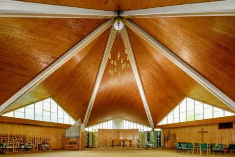 Historic England Inside view of St Richard's Church in Richmond showing the tall angular wooden roof. Crosses can be seen on the walls, as well as an altar, an organ and some chairs stacked to one side.