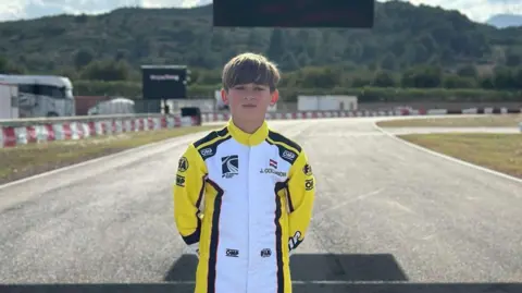 Jack Collinson, a boy with short blond hair wearing a yellow and white one-piece racing suit, is standing with the empty track in Valencia behind him. 