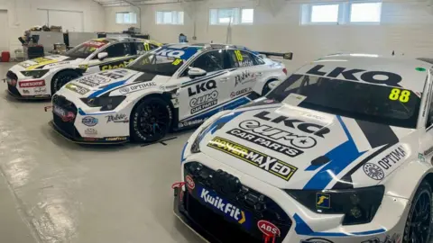Three white racing cars covered in branding are lined up in a garage with white walls and tools in the background.