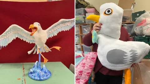 A composite of two model seagulls. The one on the left is made of paper mache. The birds wings are outstretched, and its mouth wide open, as if in flight. On the right, a large cushion is in the shape of the side profile of a seagull. There is a person wearing a red jumper who is holding up the model.