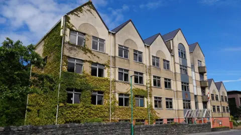 BBC A large yellow building with seven triangular points and green ivy creeping up the left side of the building. 